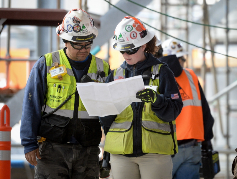 Two construction workers, members of NABTU, in hard hats and reflective vests consulting a paper document on a worksite.