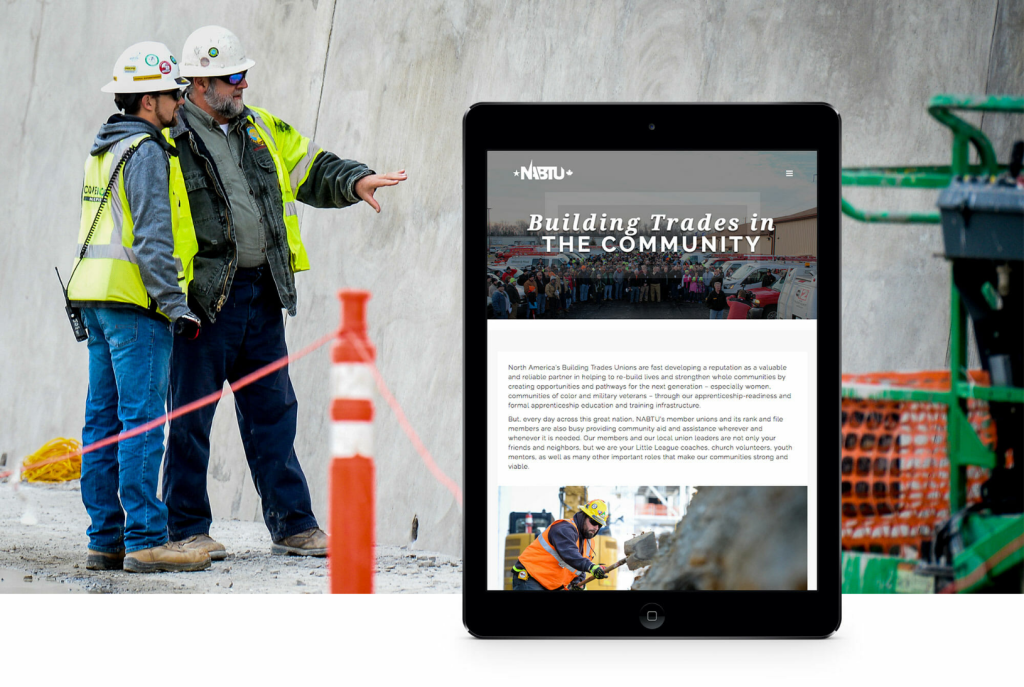 Two construction workers in hard hats engrossed in a discussion over a digital tablet with a presentation titled "NABTU building trades in the community" visible on the screen, at a construction site.