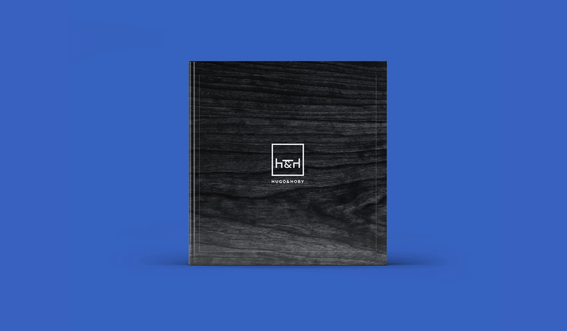 A sleek black journal with an embossed Hugo & Hoby geometric logo centered on a textured cover, against a vibrant blue background.