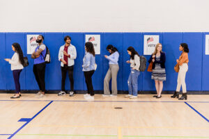 Print Design for Political Campaigns A diverse group of people standing in line against a blue wall with "vote" posters, waiting to cast their votes in a gymnasium during a political campaign.