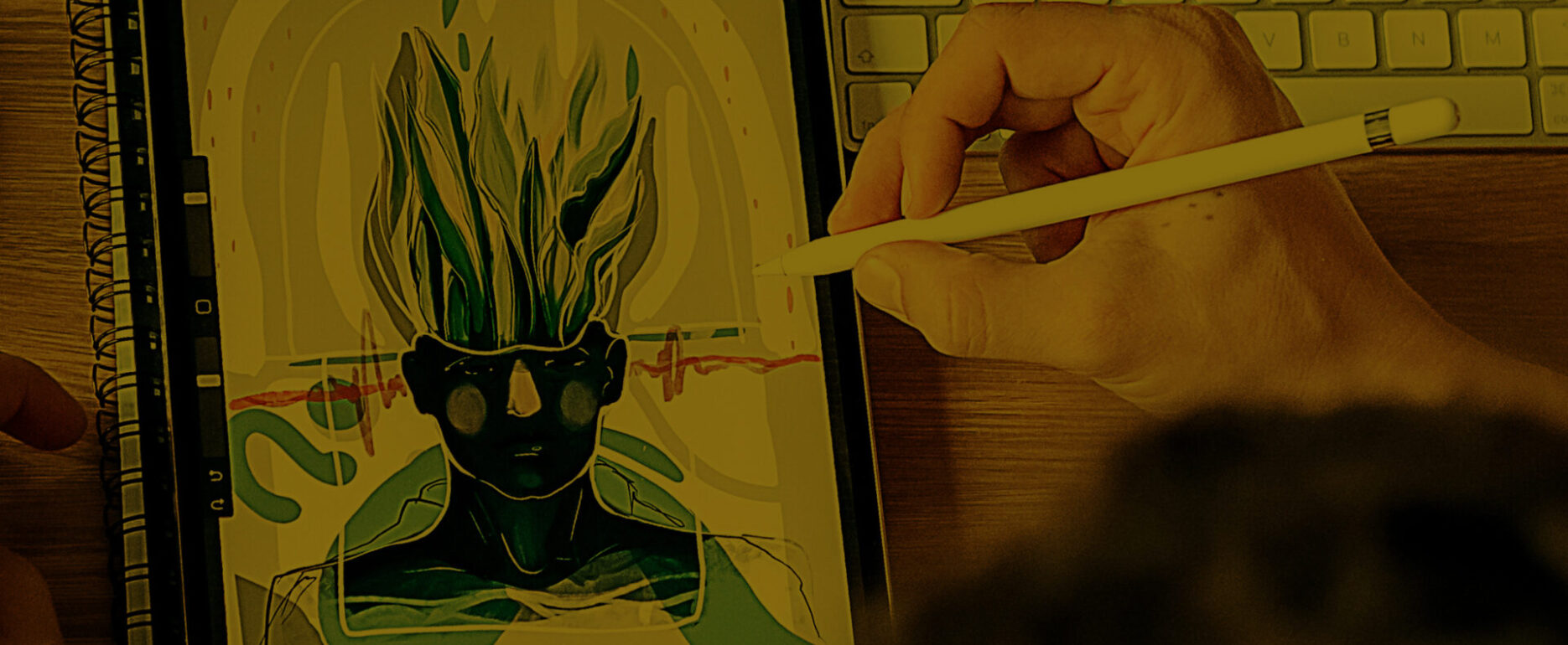 A person uses a stylus on a tablet to create digital art featuring a figure with flame-like hair and sunglasses. The tablet is placed on a wooden desk beside a sketchbook, keyboard, and an open laptop.