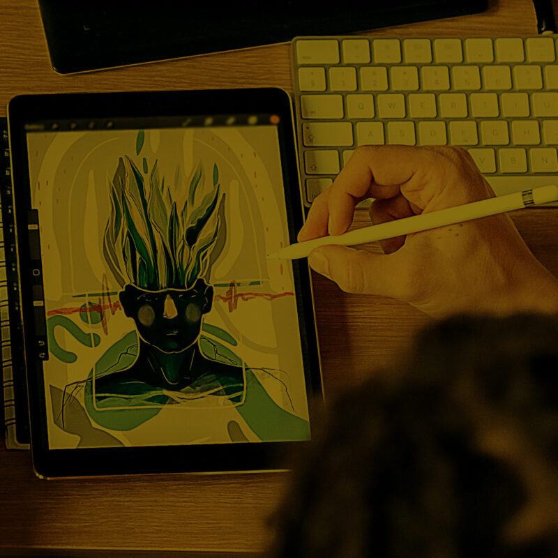 A person uses a stylus on a tablet to create digital art featuring a figure with flame-like hair and sunglasses. The tablet is placed on a wooden desk beside a sketchbook, keyboard, and an open laptop.
