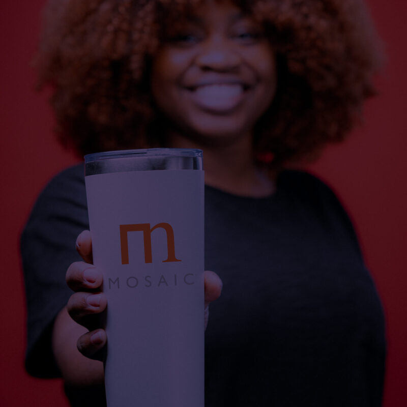A person with curly hair smiling and holding out a white tumbler with the word "MOSAIC" and an orange "M" logo on it. The background is red, and the person is wearing a dark shirt. The focus is on the tumbler, with the person slightly blurred in the background.