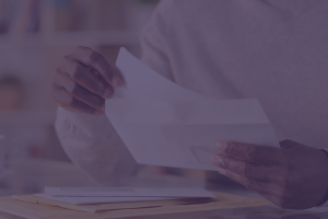 A person's hands holding an open envelope, appearing to retrieve or insert a letter. They are seated at a table with other papers and documents visible in the background, suggesting they are immersed in postage and mailing regulations. The image has a purple tint.