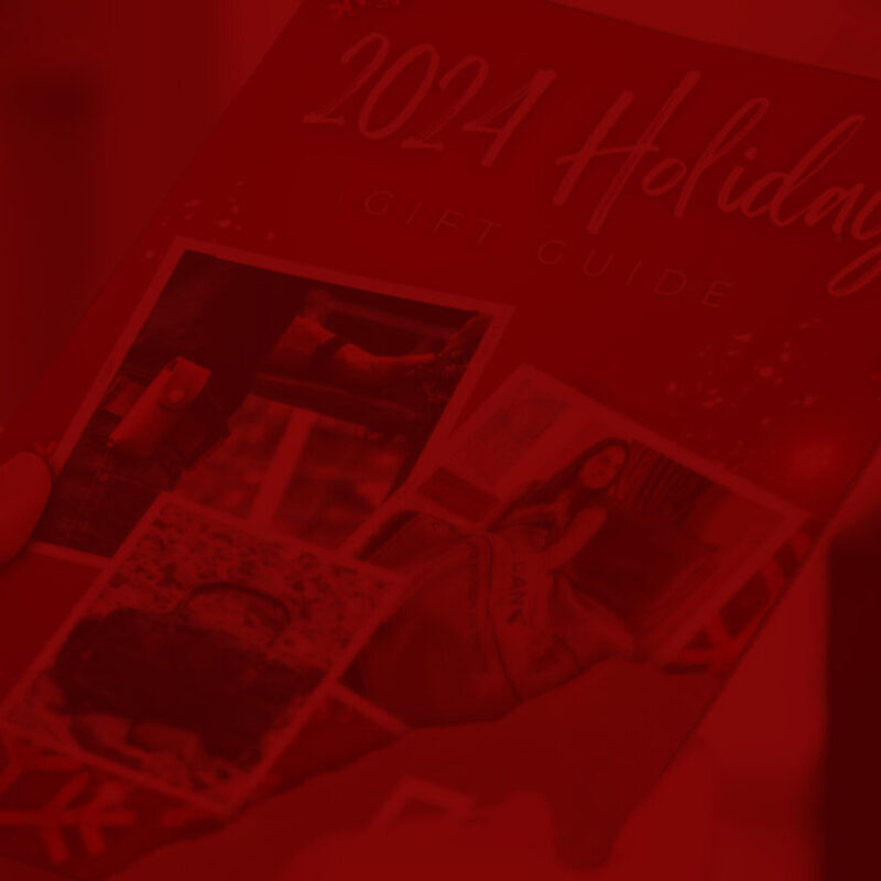 A person holds a brochure titled "2024 Holiday Gift Guide." The cover features several photos, including a close-up of a wrapped gift, a woman in festive clothing, and holiday decorations. The background is blurred, focusing attention on the brochure.