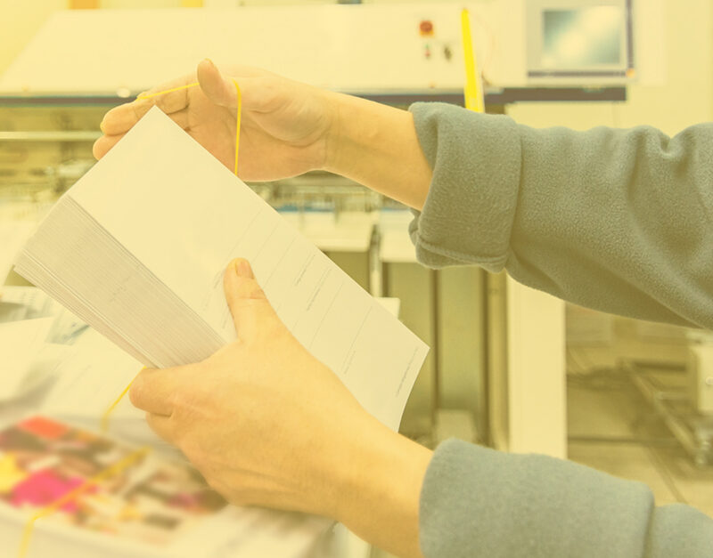 A person holds a stack of envelopes tied with a yellow rubber band in a bustling printing facility, reminiscent of the organized efficiency behind mPower Ecommerce Storefront. The background, slightly blurred, reveals the hum of additional printed materials ready to be dispatched.