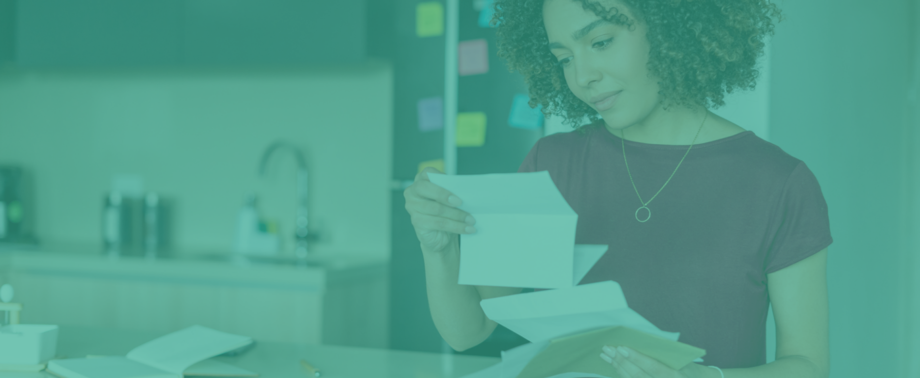 A woman with curly hair stands at a kitchen counter, analyzing direct mail trends to watch for in 2025 as she reads through several letters and envelopes. The background blurs into a teal-tinted kitchen sink and a wall peppered with sticky notes, adding an aura of contemplation.