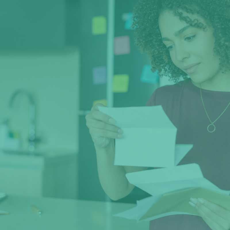 A woman with curly hair stands at a kitchen counter, analyzing direct mail trends to watch for in 2025 as she reads through several letters and envelopes. The background blurs into a teal-tinted kitchen sink and a wall peppered with sticky notes, adding an aura of contemplation.