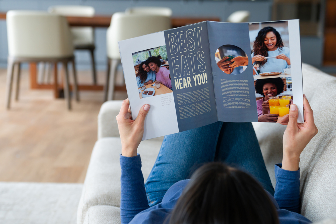 Person reading a magazine titled "Best Eats Near You!" with images of people enjoying food and drinks. The person is lying on a couch, and the room features a dining table and chairs in the background.