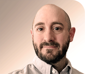 A bearded man with a shaved head is looking at the camera and smiling slightly. He is wearing a light-colored collared shirt. The background is a gradient of beige tones.