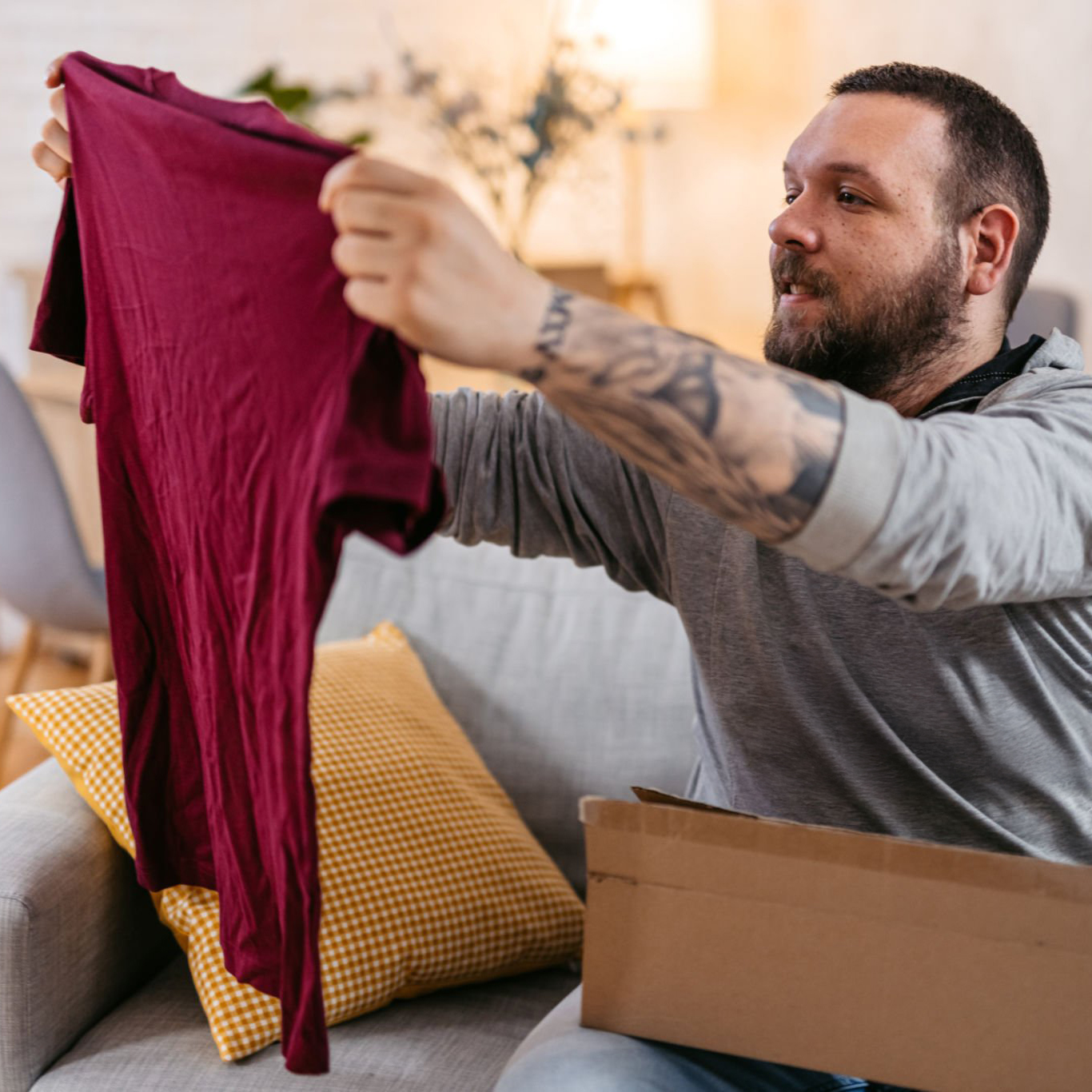 A bearded man with tattoos holds up a maroon shirt with a smile, sitting on a couch beside a cardboard box from his favorite Storefronts brand. A yellow pillow rests nearby, set against the soft-focus backdrop of his welcoming living room.