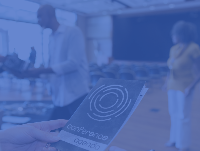 A person holds a spiral-designed conference agenda, titled "Creative Resolutions: How to Reimagine Your Marketing Strategy in 2025." In the spacious room behind, people casually interact amidst rows of chairs. The scene is subtly enhanced by a blue tint overlay.