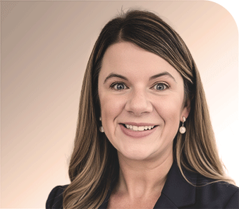 Woman with long brown hair smiling at the camera, wearing a dark blazer and pearl earrings, in front of a neutral gradient background.