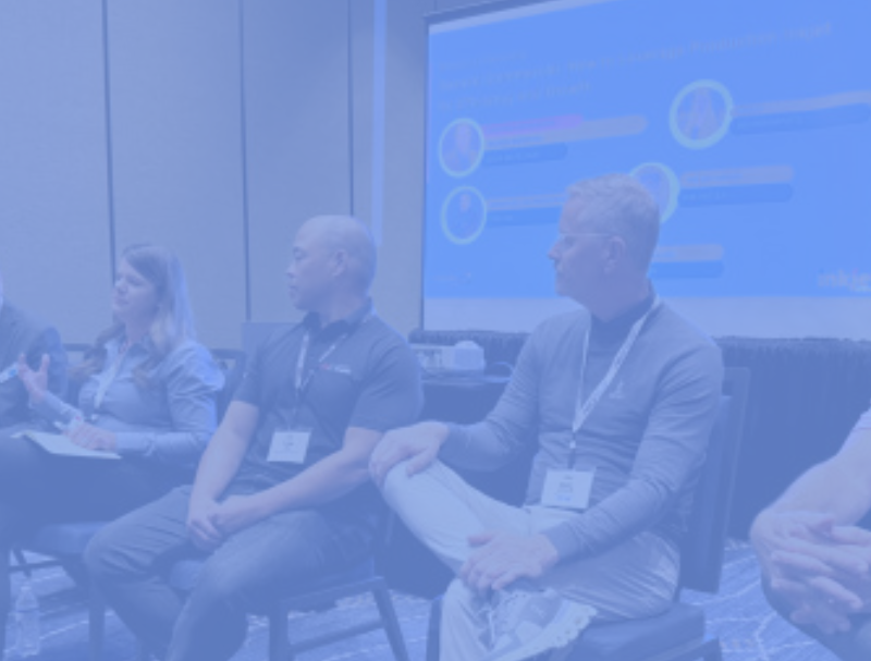Five people sit on chairs in a conference room, participating in a panel discussion. One woman holds a microphone while speaking about "Inside the 2025 Inkjet Summit: MOSAIC's Perspective on What’s Next," with a projection screen displaying graphics behind them.