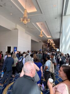 A large crowd gathers in a brightly lit hallway at the BRIDGE Conference, with many marketing teams talking, holding bags, or carrying coffee. Chandeliers hang from the high ceiling above.