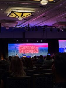 A conference room audience at the BRIDGE Conference faces a stage with a large screen displaying "IGNITED BY PURPOSE" in bold red letters over a city skyline. The dimly lit room and decorative ceiling lights inspire marketing teams and fundraising professionals alike.