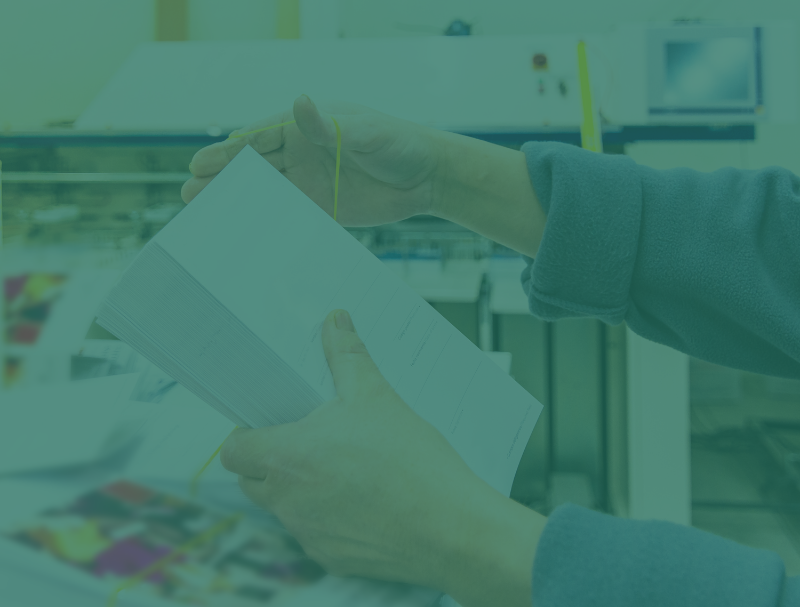 A person holds a stack of printed papers tied with a yellow string in a print shop, surrounded by printing equipment and materials—perfect for Marketing & Fundraising campaigns.