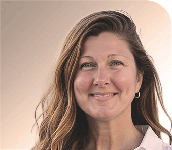 Smiling woman with long wavy brown hair, wearing a white shirt and small hoop earring, standing in front of a soft beige background.